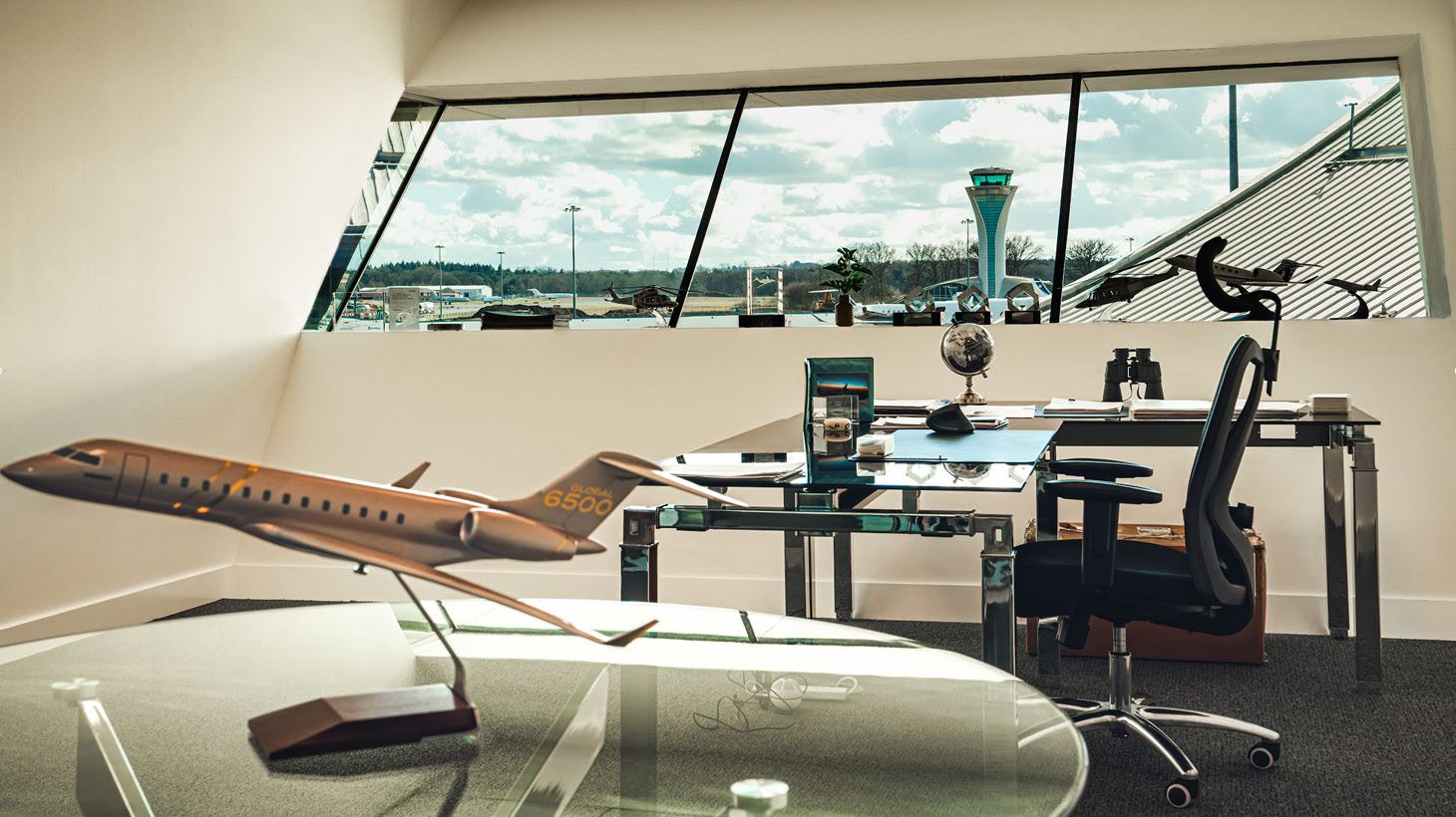 a glance in the office shows a bronze model aircraft on a glass table, an elegant desk and a beautiful view outside the windows on the airstrip